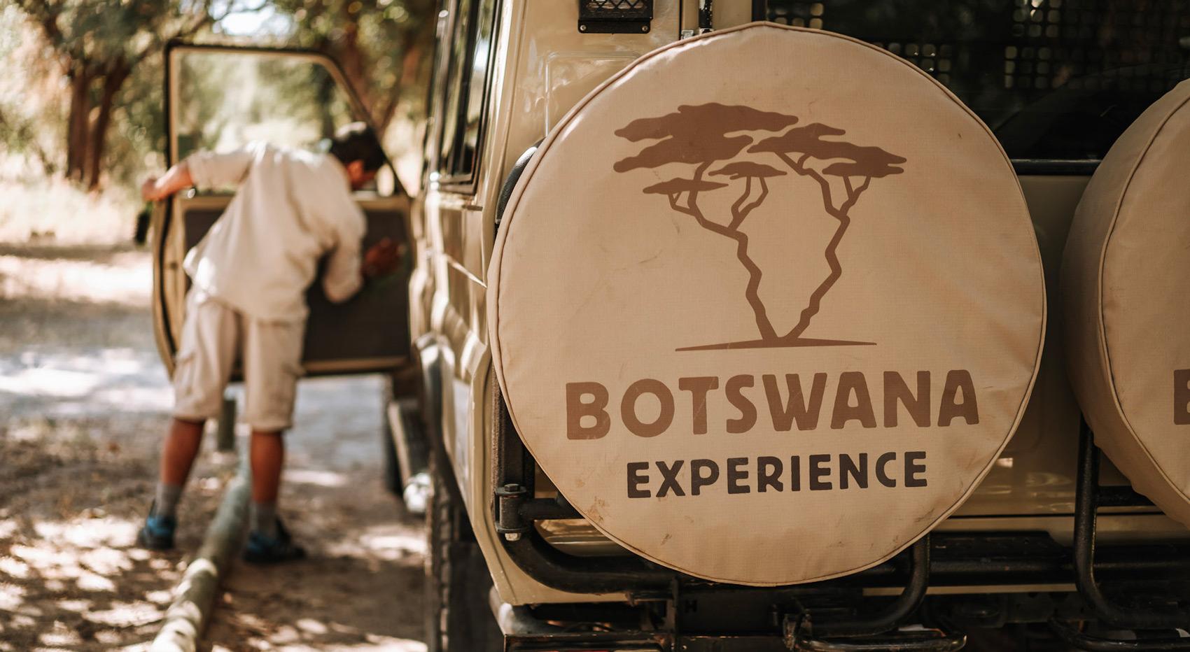 A photo of the tirecover on a the Botswana Experience vehicle with a guide in the background. A photo of the tirecover on a the Botswana Experience vehicle with a guide in the background.